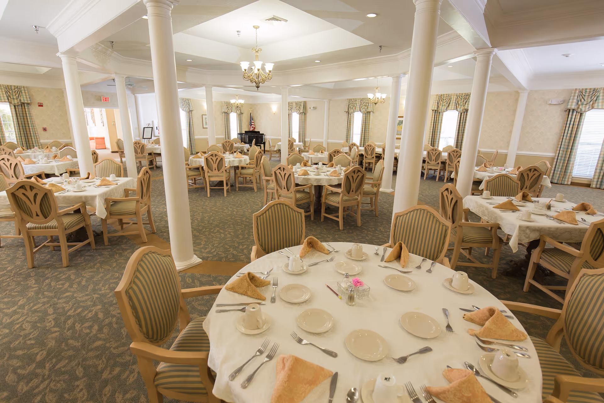 Large, well-lit dining room with multiple round tables neatly set with dishes, utensils, and folded napkins.