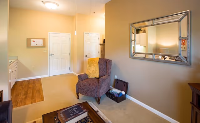 A cozy living room area with beige walls and carpet. There is a patterned armchair with a yellow throw blanket, a wooden coffee table with books on it, and a large decorative mirror on the wall reflecting part of the room. Two white doors are visible in the background along with a small section of a kitchen area with white cabinets and wooden flooring.