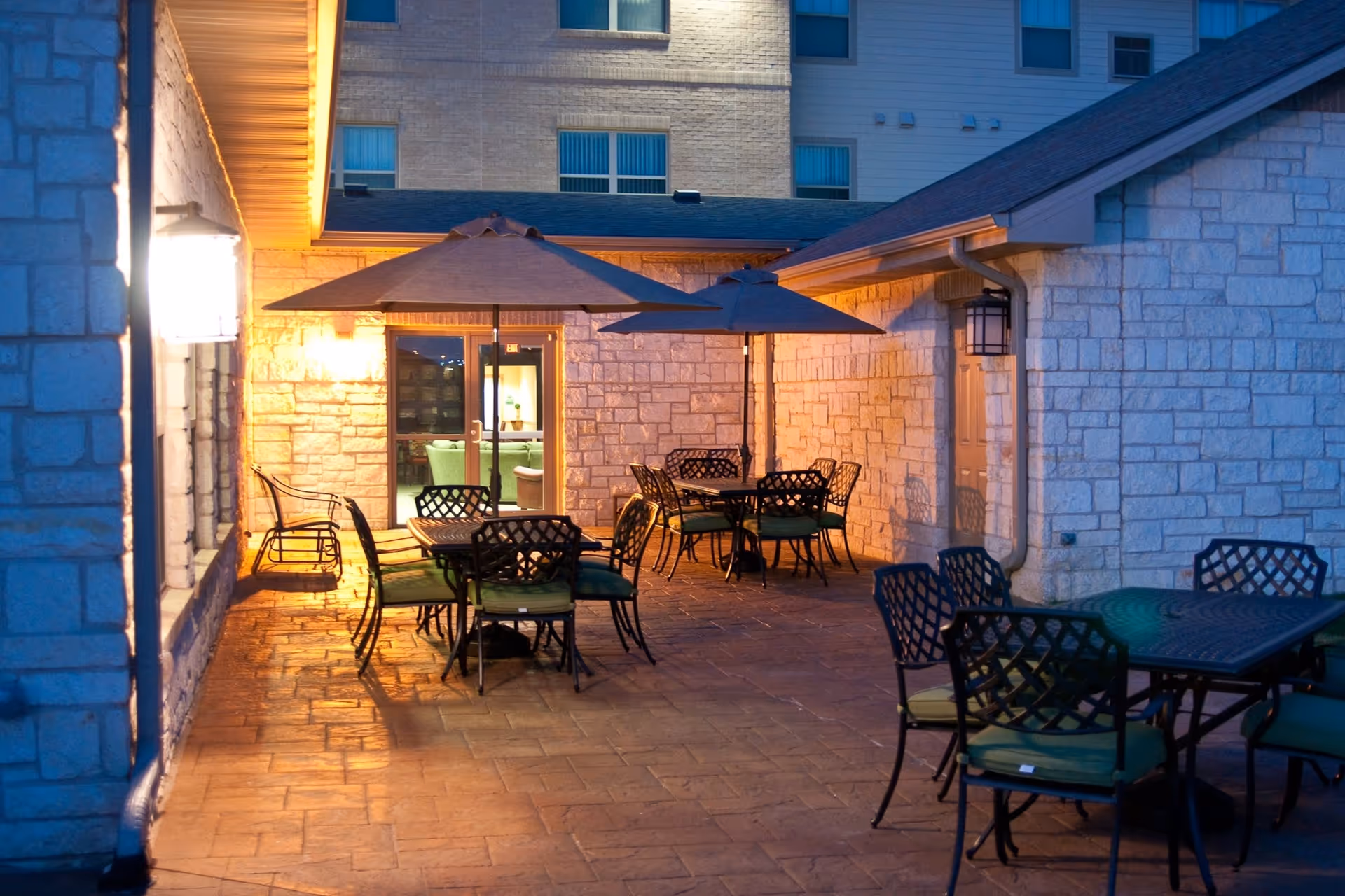 Outdoor patio area at dusk with several tables and chairs, each table shaded by large umbrellas. The patio is surrounded by stone walls and is softly lit by wall-mounted lights. A glass door leads inside the building, where more seating is visible.