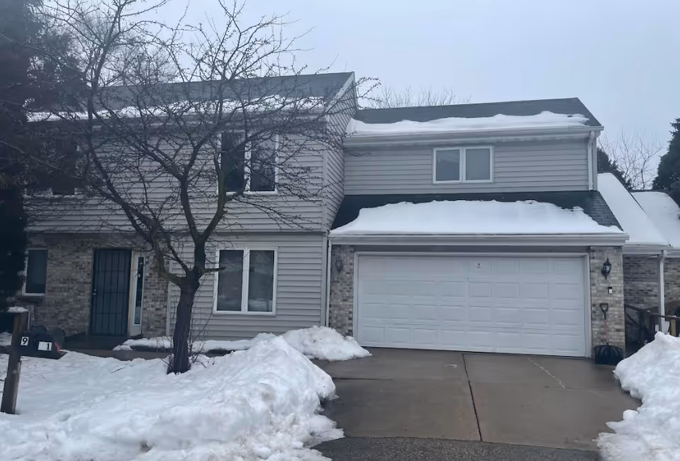Two-story townhouse with an attached two-car garage, a bare tree in front, and snow on the roof and ground.