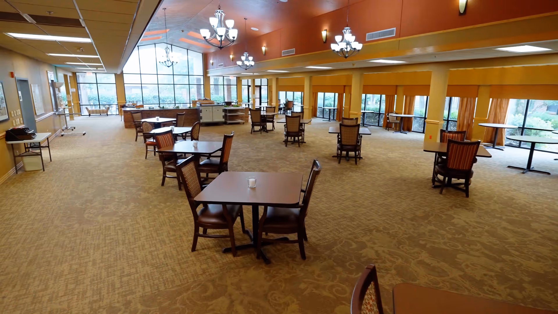 Spacious dining area with multiple tables and chairs arranged throughout the room. Large windows along the walls allow natural light to fill the space. The ceiling features several chandeliers and recessed lighting. The room has a warm color palette with beige carpet and orange walls.