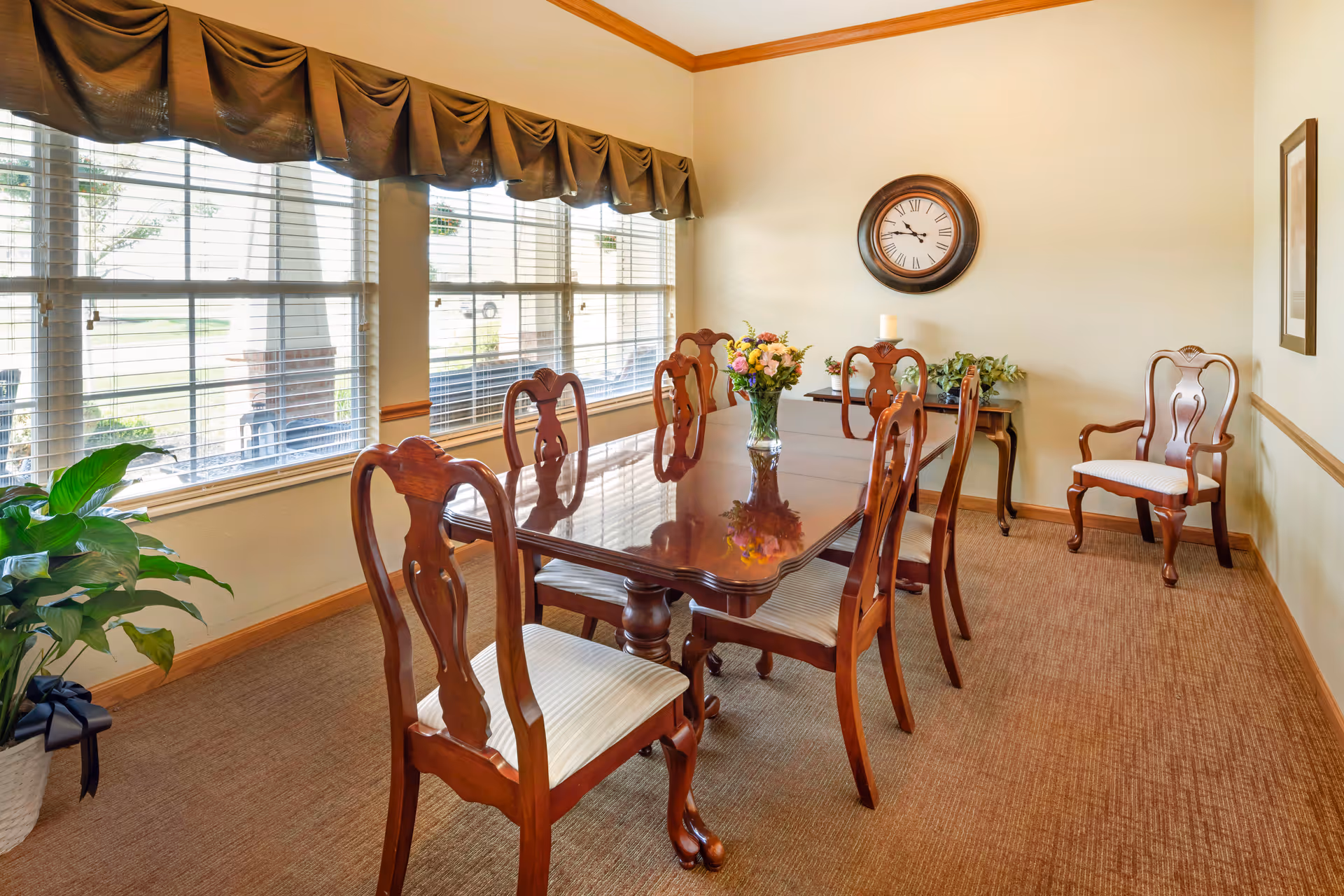 A bright dining room with a polished wooden table surrounded by six matching wooden chairs with white cushions. A vase with colorful flowers is placed in the center of the table. Large windows with blinds and brown valances let in natural light. A wall clock with Roman numerals hangs on the beige wall, and a small table with plants and a candle is in the corner. A potted plant with a black ribbon is on the floor near the windows.