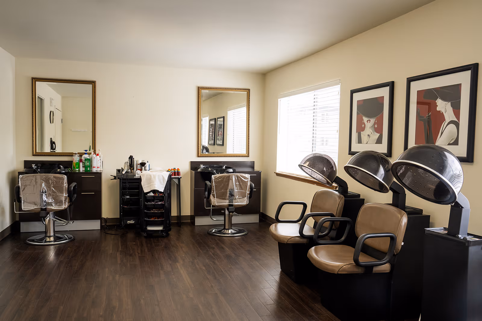 Interior view of a hair salon area with two styling chairs in front of mirrors and two hair drying chairs with hooded dryers. The room has wooden flooring, light-colored walls, and two framed artworks on the wall near a window with blinds.