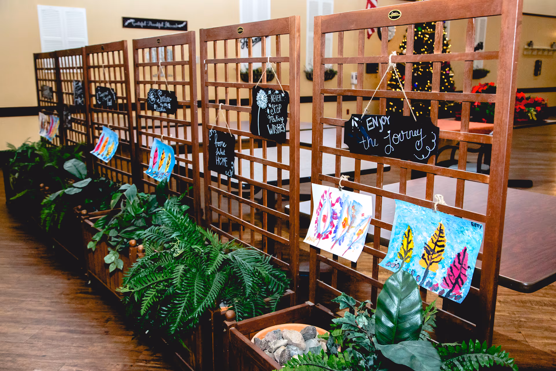 Wooden lattice room dividers decorated with small chalkboard signs featuring inspirational quotes and colorful paintings, with green plants in wooden planters at the base, set in a room with wooden flooring and a decorated Christmas tree in the background.
