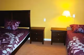A bedroom with two single beds separated by two wooden nightstands. Each bed has a purple bedspread with a white leaf pattern and a matching pillow. A wall-mounted lamp is above the right nightstand, illuminating the yellow wall behind.