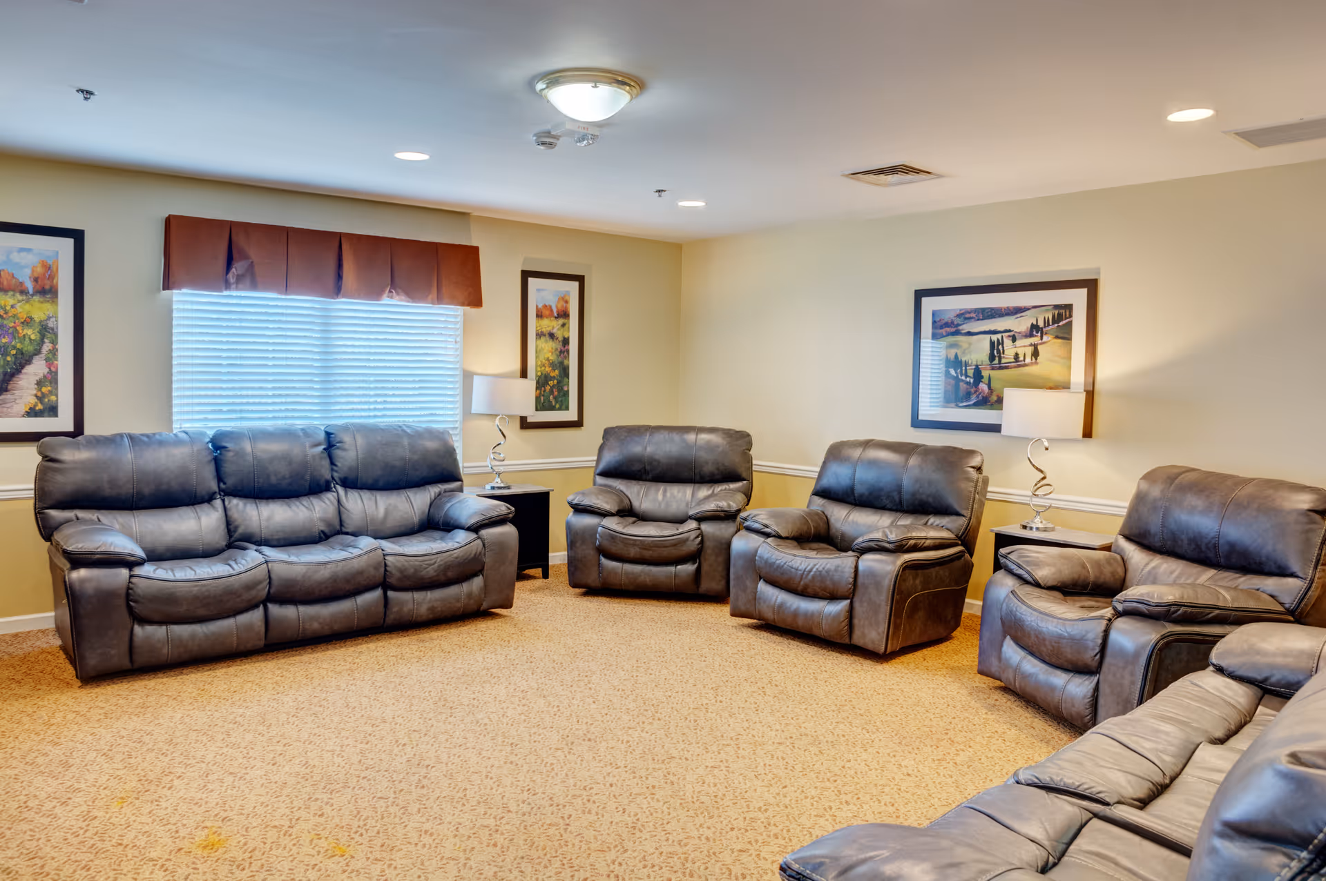 A cozy living room with multiple dark leather recliner sofas and armchairs arranged in a semi-circle on a beige carpet. The walls are light yellow with three framed landscape paintings and two side tables with modern lamps. A window with blinds and a brown valance is visible on the left wall.