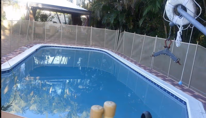 Rectangular backyard pool enclosed by beige safety fence with trees and shade structure.