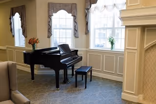 A cozy interior room featuring a black grand piano with a matching bench, positioned near three large windows with decorative curtains. A vase with flowers is placed on the piano and another vase with flowers is on the windowsill. The room has beige walls with white wainscoting and a fireplace on the right side.