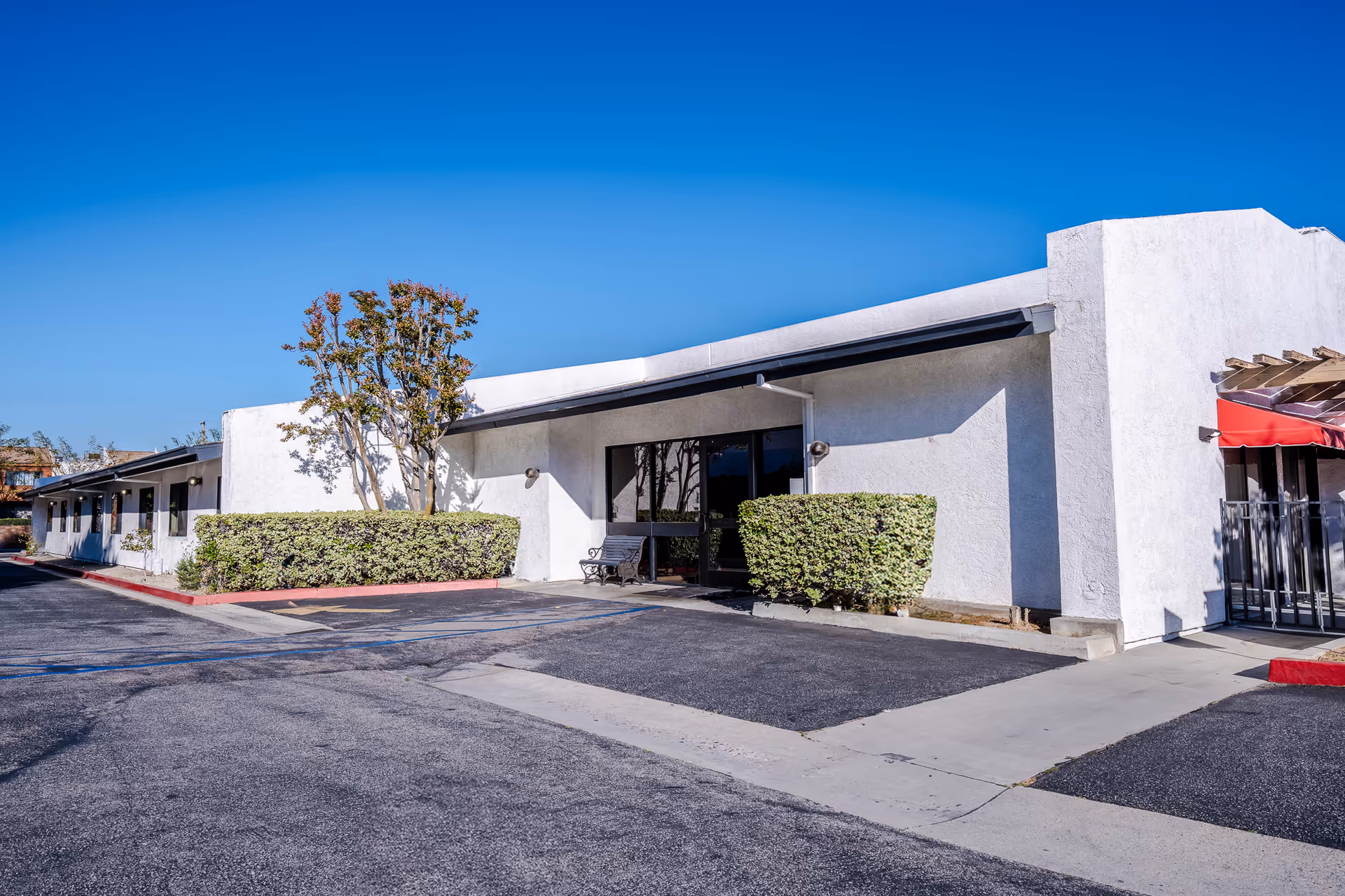Exterior view of a single-story white building with a flat roof, surrounded by trimmed bushes and a tree. There is a bench near the entrance with glass doors, and a red awning is visible on the right side. The sky is clear and blue.