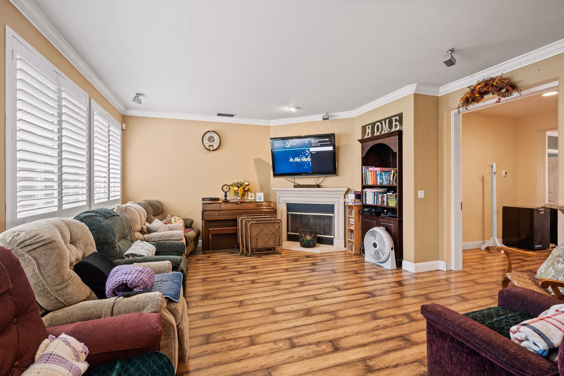 Spacious living room with several upholstered recliners around a fireplace and wall-mounted TV, wood floors, bookshelf, and large shuttered windows.
