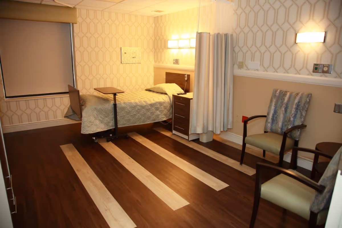 A nursing facility room with a single bed covered in a light quilt, a small overbed table, a nightstand, two cushioned chairs, and a privacy curtain. The room has patterned wallpaper, wood flooring with light stripes, and soft lighting fixtures on the walls.
