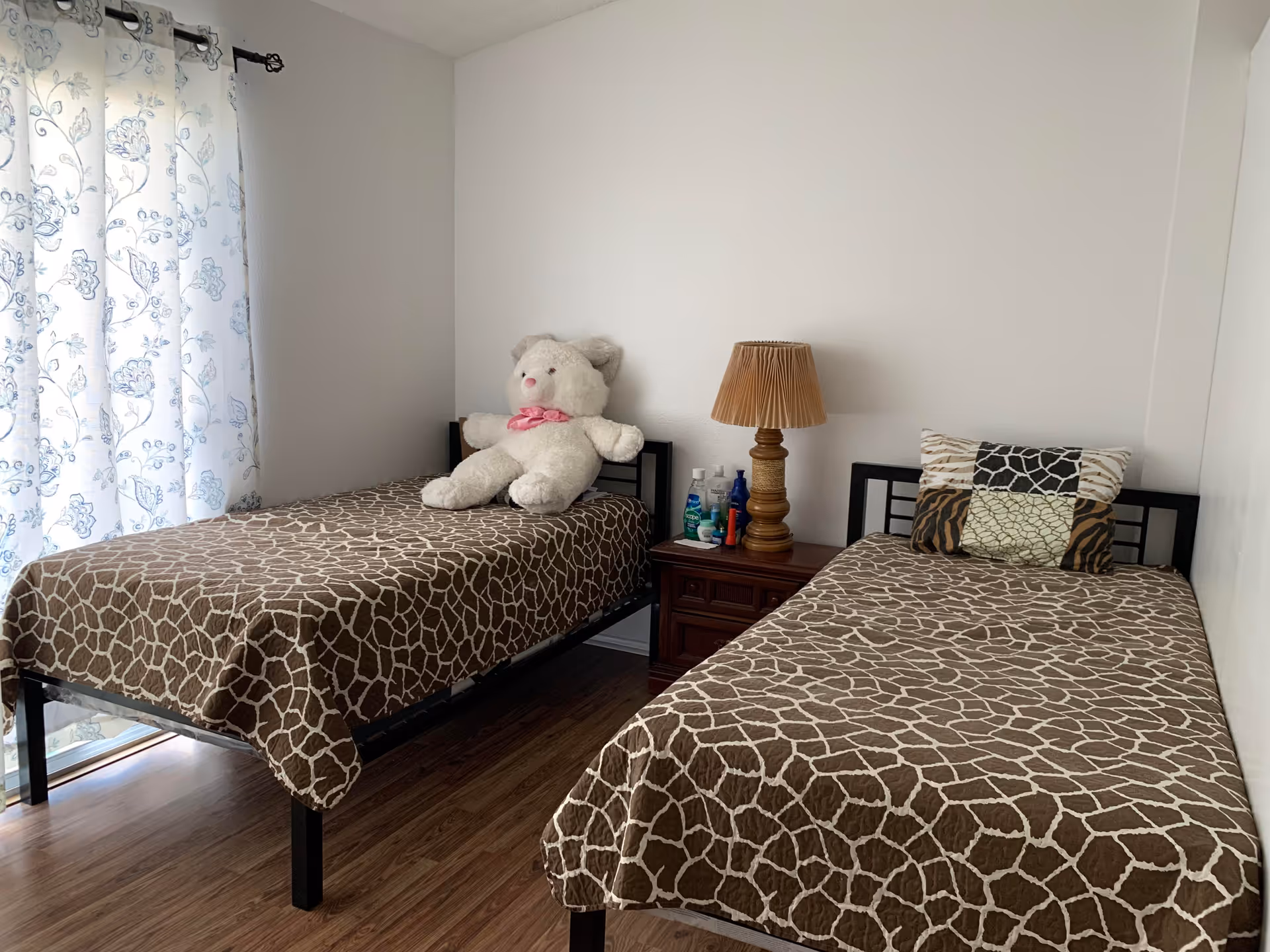 Small bedroom with two twin beds covered in giraffe-print bedspreads, a nightstand with a lamp and toiletries, and a large teddy bear on one bed.