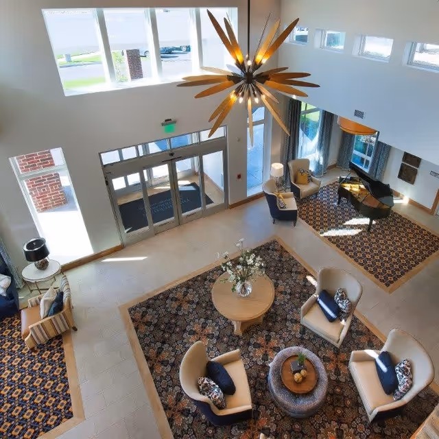 A bright and spacious senior living facility lobby with large windows allowing natural light to fill the room. The area features a modern chandelier, patterned rugs, comfortable armchairs with decorative pillows, a round table with a floral arrangement, a grand piano, and a glass entrance door.