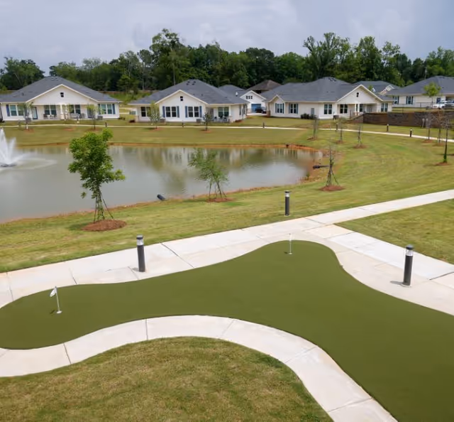 Outdoor view of a senior living facility featuring a small pond with a fountain, surrounded by green lawns and young trees. In the foreground, there is a putting green with two golf holes and a paved walkway. Several single-story residential buildings with gray roofs are visible in the background under a partly cloudy sky.