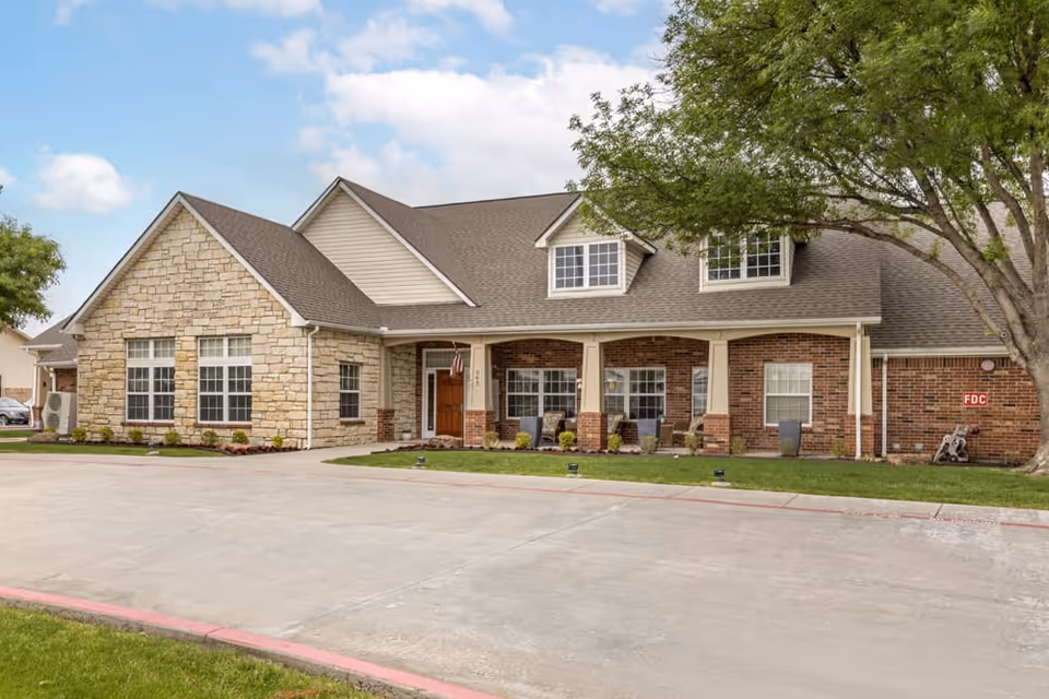 Exterior view of a single-story senior living facility building with a combination of stone and brick facade, a covered porch with seating, and a large tree on the right side. The driveway and lawn are visible in the foreground under a partly cloudy sky.