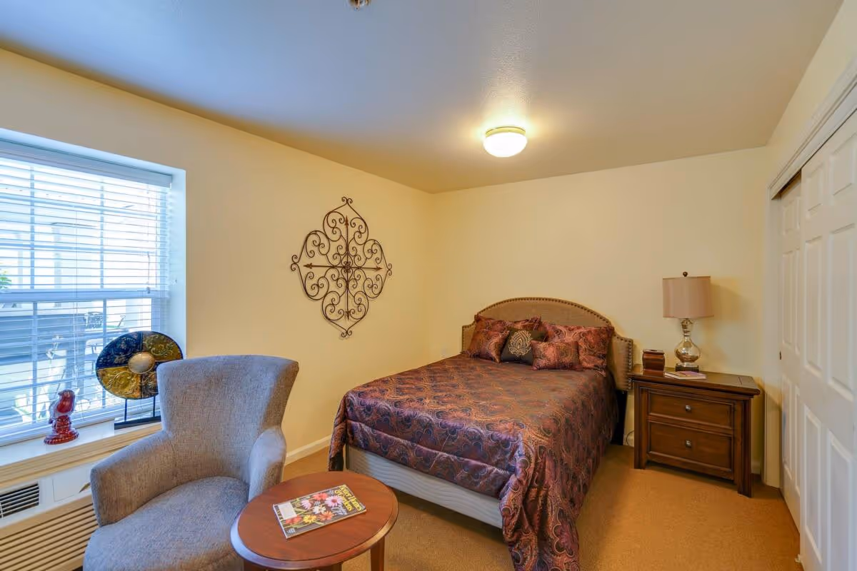 A cozy bedroom with a double bed covered in a patterned bedspread and multiple pillows. There is a wooden nightstand with a lamp and some books next to the bed. A comfortable gray armchair and a small round wooden table with a magazine are placed near a window with white blinds. The walls are light-colored with a decorative metal wall hanging above the bed.