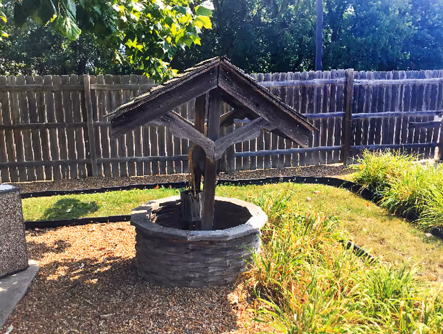 A decorative wooden wishing well sits in a gravel garden bed surrounded by plants and a wooden fence.