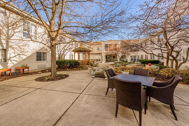 Outdoor courtyard area at Eden Vista Burr Ridge featuring a paved patio with a table and four wicker chairs, leafless trees, a wooden gazebo, and surrounding two-story buildings under a clear blue sky.