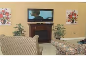 A cozy living room area with a flat-screen TV mounted above a wooden fireplace. There are two armchairs, one with a patterned fabric and the other floral, positioned facing the TV. Two potted plants flank the fireplace, and floral paintings hang on the beige walls.
