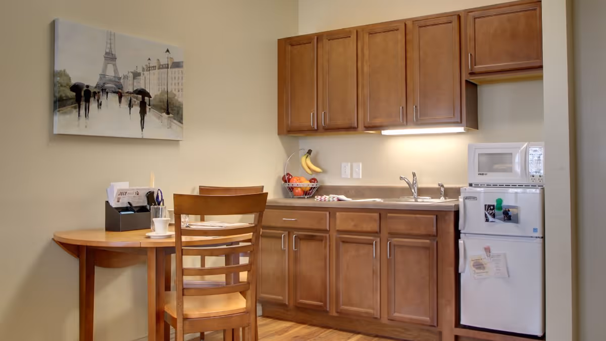 A small kitchen area with wooden cabinets, a countertop with a sink, a microwave on top of a mini refrigerator, and a wooden table with two chairs. A fruit basket with bananas and apples is on the counter, and a painting of people walking near the Eiffel Tower hangs on the wall.