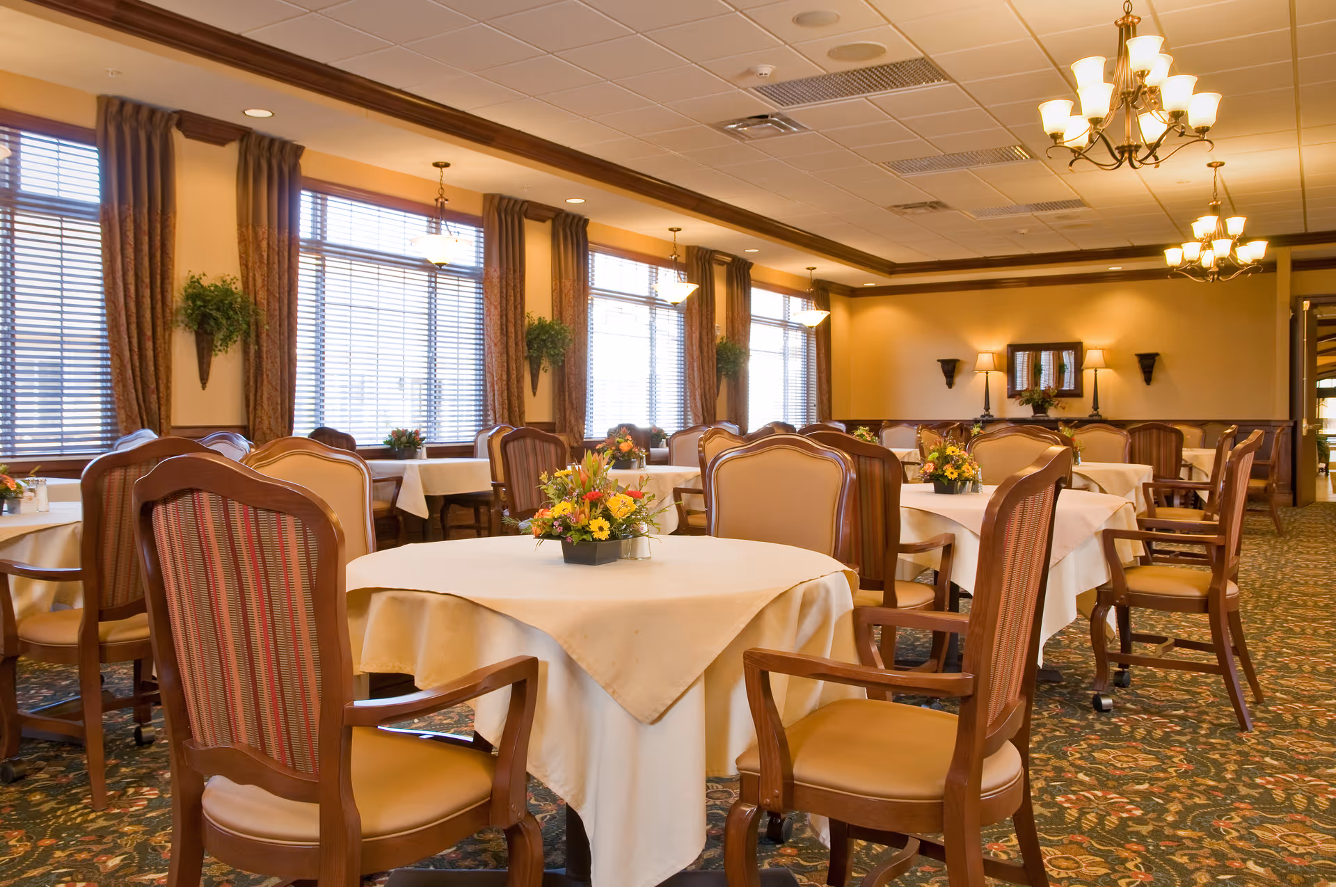 A well-lit dining room with multiple round tables covered with white tablecloths, each adorned with a floral centerpiece. The room features wooden chairs with cushioned seats and backs, large windows with blinds and curtains, wall-mounted plants, and warm lighting from chandeliers and wall sconces. The carpet has a floral pattern, and the overall ambiance is cozy and inviting.