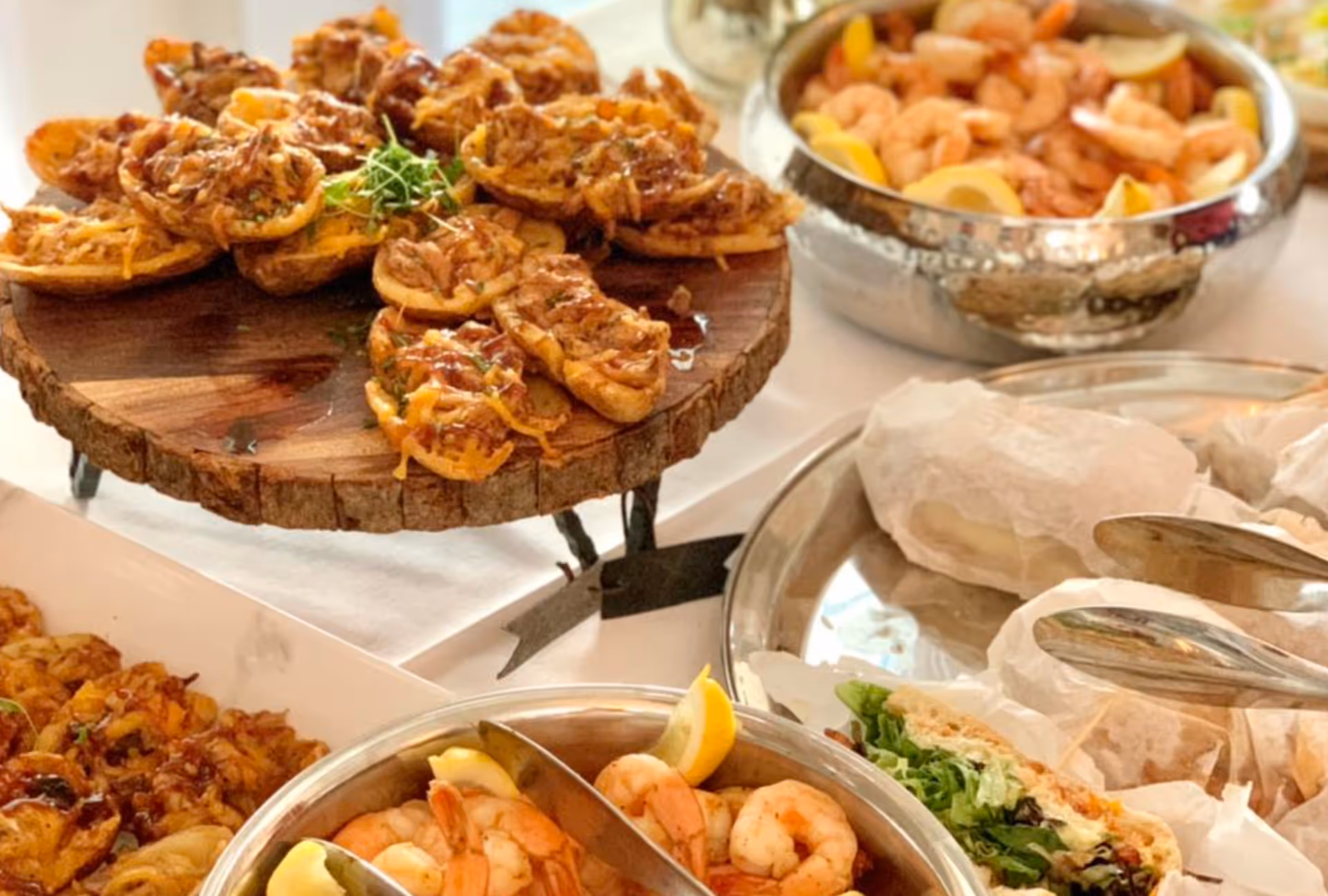 A close-up view of a buffet table featuring a wooden stand with small baked appetizers topped with melted cheese and herbs, a metal bowl filled with shrimp garnished with lemon wedges, and wrapped sandwiches with lettuce and other fillings on a silver tray.