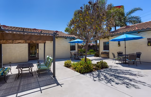Outdoor courtyard area with concrete flooring, a small tree surrounded by greenery in the center, patio tables with blue umbrellas, and cushioned chairs under a shaded pergola. The buildings have beige stucco walls and red tile roofs under a clear blue sky.