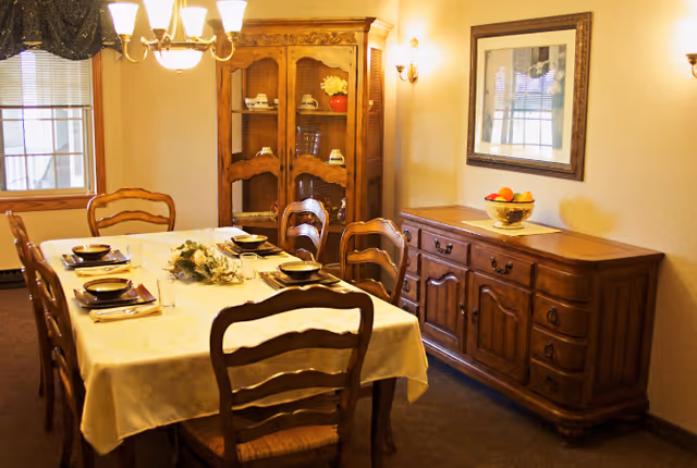 A warmly lit dining room with a rectangular table covered with a white tablecloth, set with plates, bowls, glasses, and napkins. Six wooden chairs surround the table. In the background, there is a wooden china cabinet displaying dishes and a wooden sideboard with a bowl of fruit on top. A framed picture hangs on the wall above the sideboard, and a window with blinds and a valance is visible on the left side.