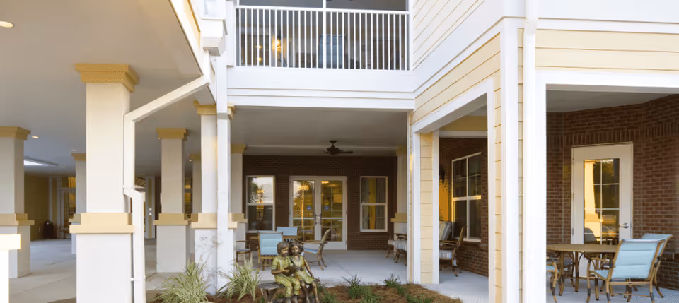 Covered outdoor courtyard with white columns, patio chairs and tables, a second-floor balcony, and a small bronze children’s statue.
