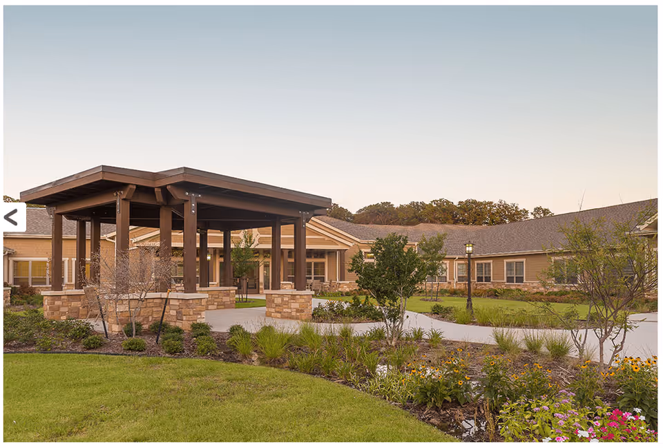 Outdoor view of a senior living facility with a covered wooden pavilion supported by stone pillars, surrounded by landscaped gardens with various plants and flowers, and a building with multiple windows in the background under a clear sky.