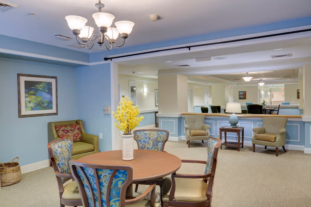 A cozy common area in a senior living facility featuring a round wooden table with a vase of yellow flowers, surrounded by four upholstered chairs with patterned backs. In the background, there are two armchairs with a small table and lamp between them, light blue walls with white trim, a framed painting, and a chandelier hanging from the ceiling.