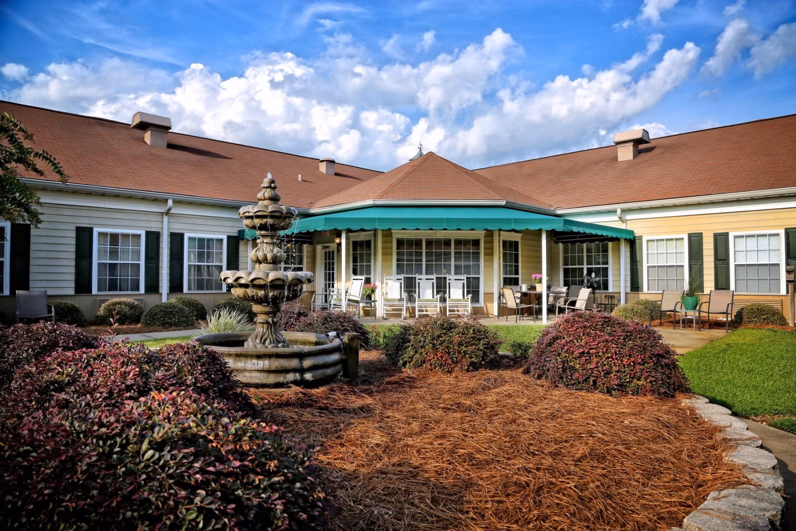 Front courtyard of a single-story senior care building featuring a tiered fountain, landscaped shrubs, and a covered patio with chairs.
