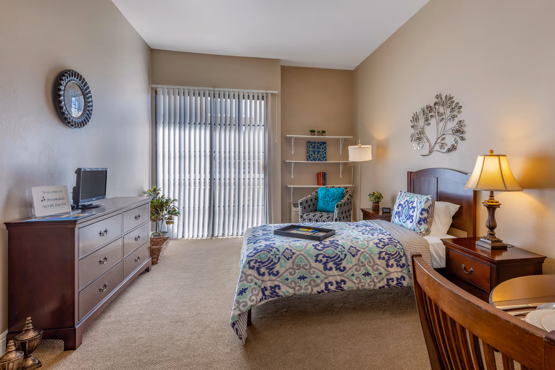 Cozy furnished bedroom with a patterned bed, wooden dresser and nightstand, armchair, and vertical blinds over a sliding glass door.