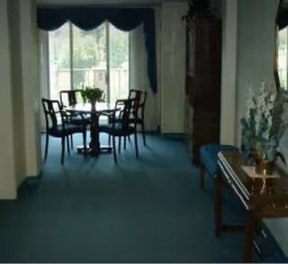 Interior view of a senior living facility showing a hallway with a small round table and four chairs near a large window with curtains. There is a wooden console table with decorative items and a bench along the right wall.