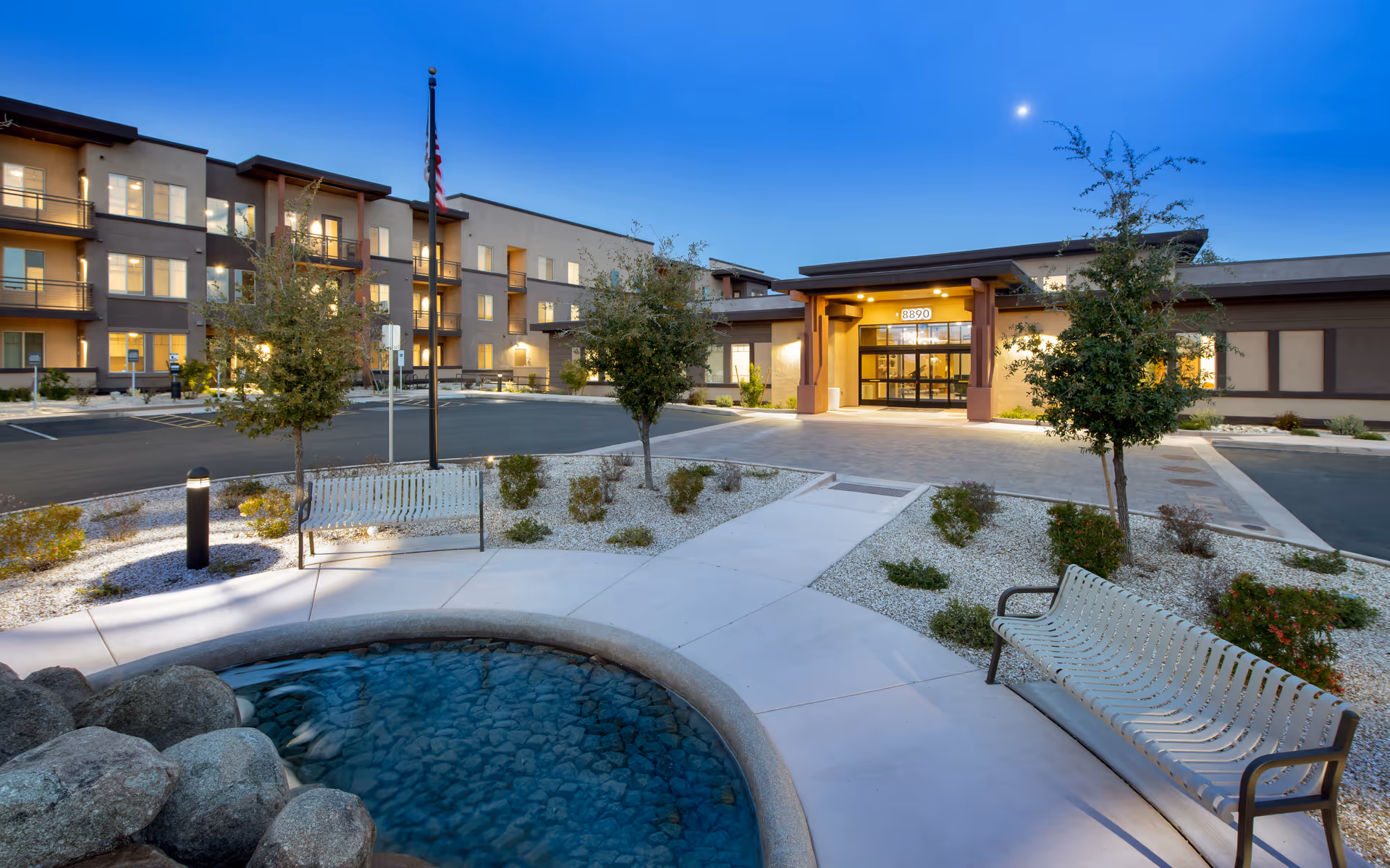 Exterior view of Revel McDowell Mountain senior living facility at dusk, showing a well-lit entrance with the address number 8890 above the door. The foreground features a small circular water feature surrounded by a paved walkway, two metal benches, small trees, and landscaped gravel areas. The building has multiple floors with balconies and windows illuminated from inside.