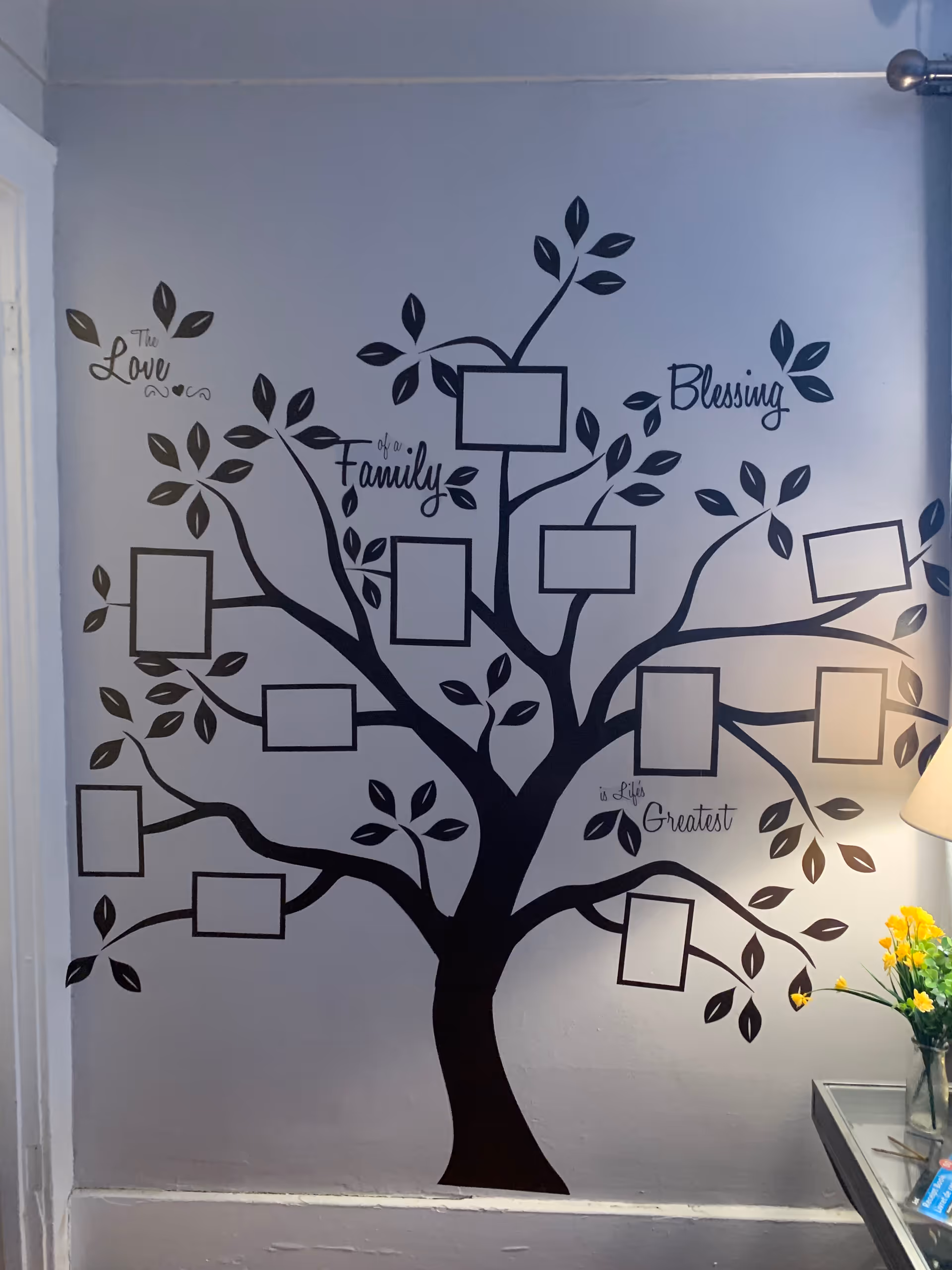 A black family-tree wall decal with blank photo frames and words like 'Family' and 'Blessing' on a pale interior wall beside a lamp and a vase of yellow flowers.