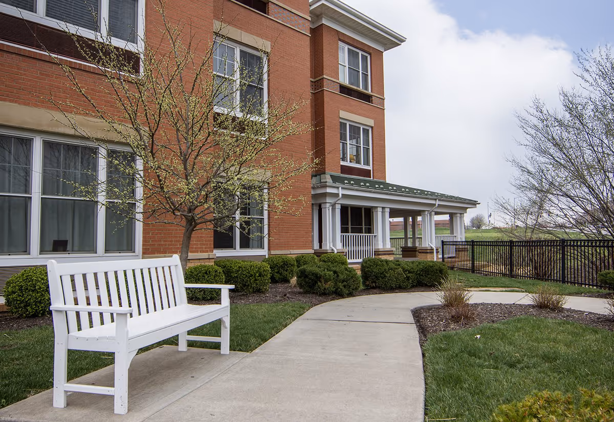 Outdoor view of a senior living facility with a white bench on a concrete pathway, small trees and bushes, and a red brick building with multiple windows in the background under a cloudy sky.