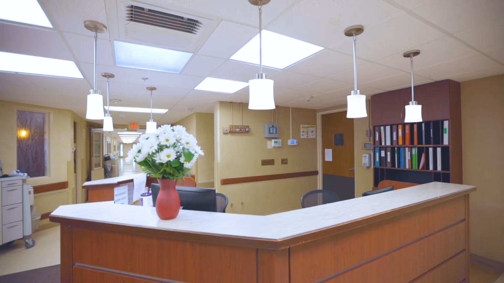 Reception desk area in a senior living facility with a vase of white flowers on the counter, overhead pendant lights, office chairs, and shelves with binders in the background. A hallway extends beyond the reception area.