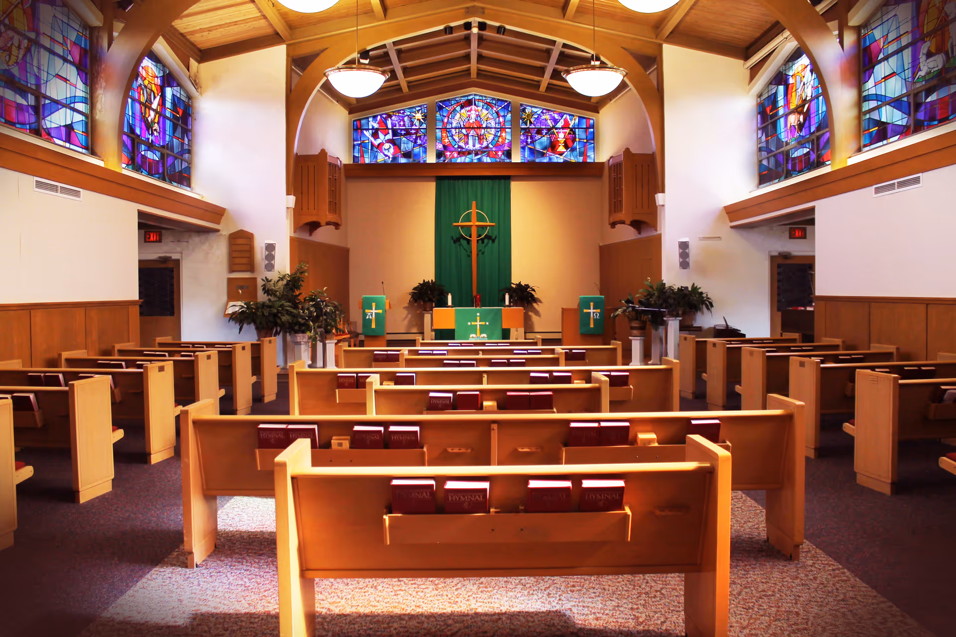 Interior view of a chapel with wooden pews, stained glass windows, and a cross at the front altar area. The ceiling is wooden with beams and hanging light fixtures.