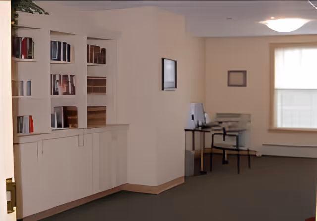 Bright common room with built-in bookshelves on the left and a small desk and chairs near a window.