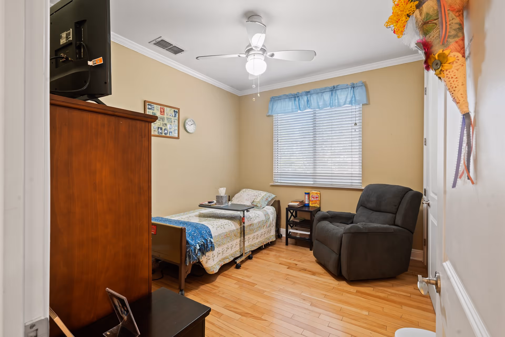 A cozy bedroom featuring a bed with a colorful quilt, a recliner chair, a small table with items, and a window with blinds.