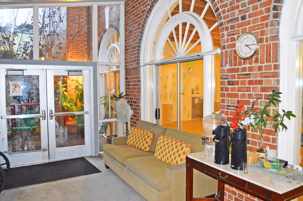 Indoor seating area with a green couch adorned with two patterned cushions, positioned against a brick wall with large arched windows. A wooden table next to the couch holds two black thermoses, a water dispenser, and some plants. Double glass doors lead outside, and a clock is mounted on the brick wall above the table.