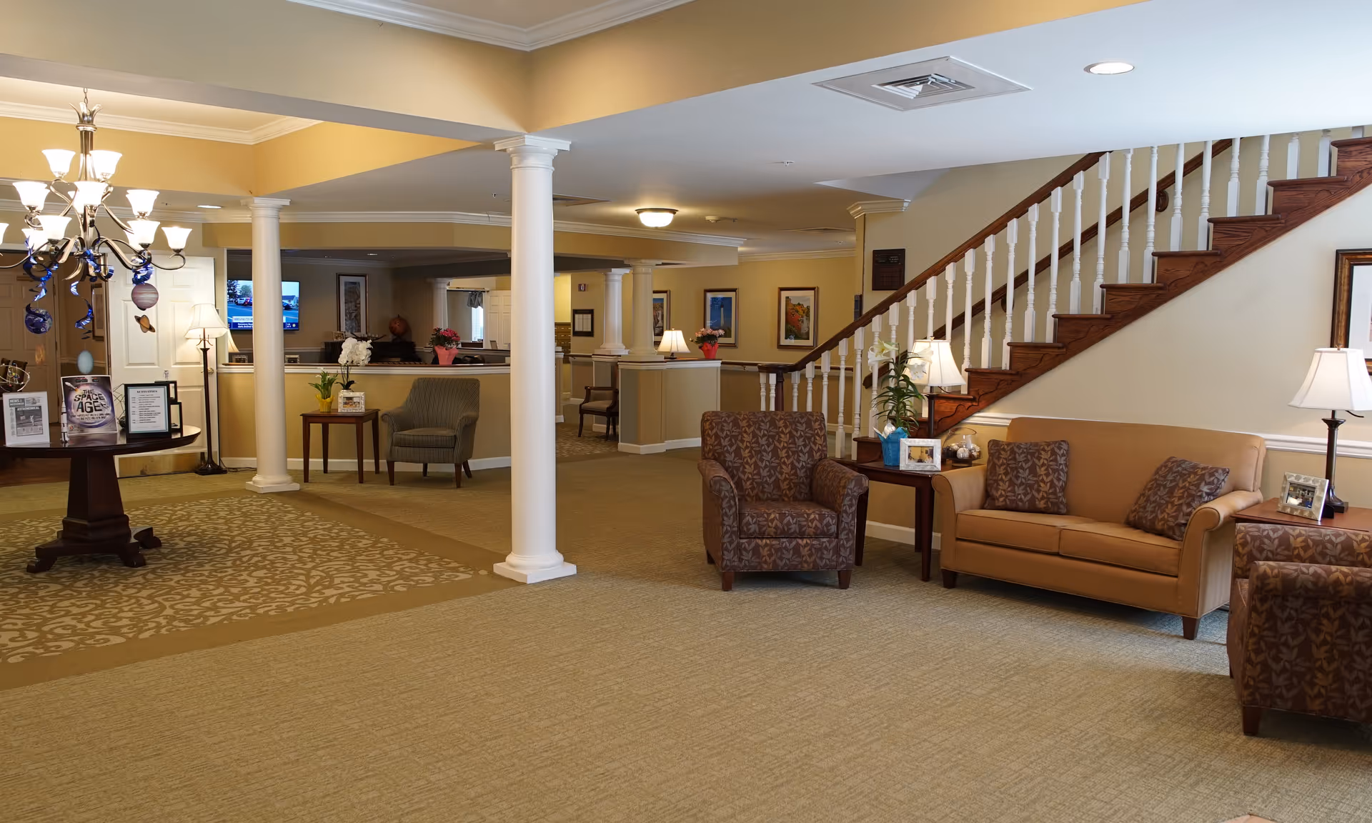 A spacious and well-lit senior living facility common area with beige walls and carpet. The room features a brown sofa with patterned cushions, two matching armchairs, side tables with lamps and framed photos, a staircase with wooden steps and white railings, white columns, a round table with informational materials and a chandelier overhead. The background shows a hallway with more seating and framed artwork on the walls.