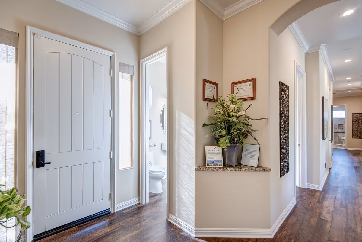 Interior view of a hallway in a senior living facility with a white front door on the left, a small bathroom visible through an open door, a corner shelf with a flower arrangement and framed certificates, and a long corridor with wooden flooring and recessed ceiling lights.