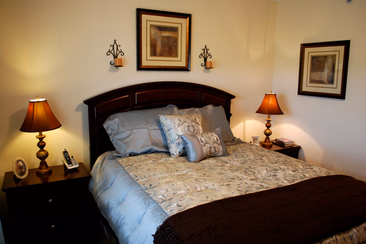 Well-made bed with decorative pillows and a dark wood headboard flanked by two bedside tables and lamps, with framed artwork on the walls.