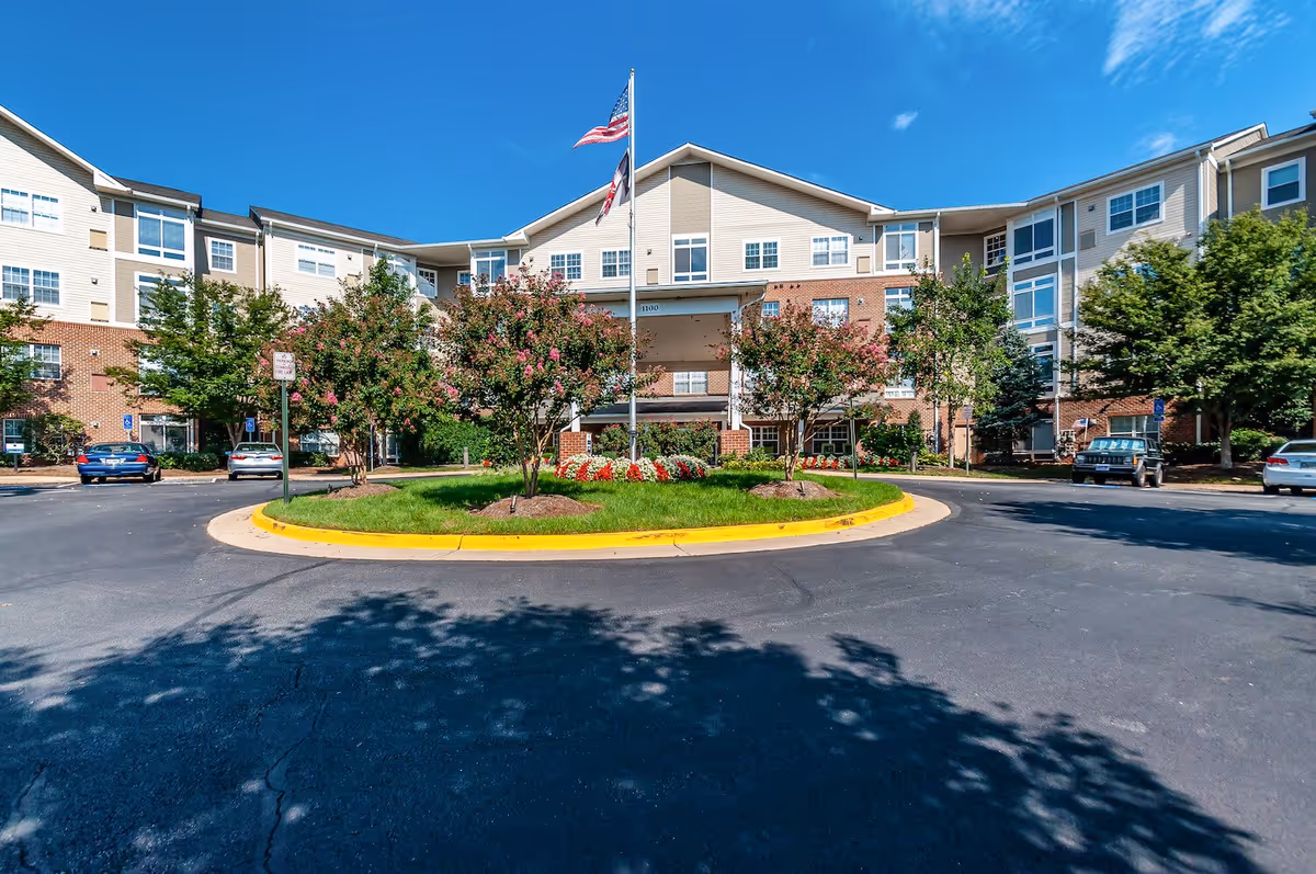 Front exterior view of Arbor Terrace Herndon, a multi-story senior living facility with beige and brick facade, surrounded by trees and a circular driveway with a flagpole displaying the American flag and another flag.