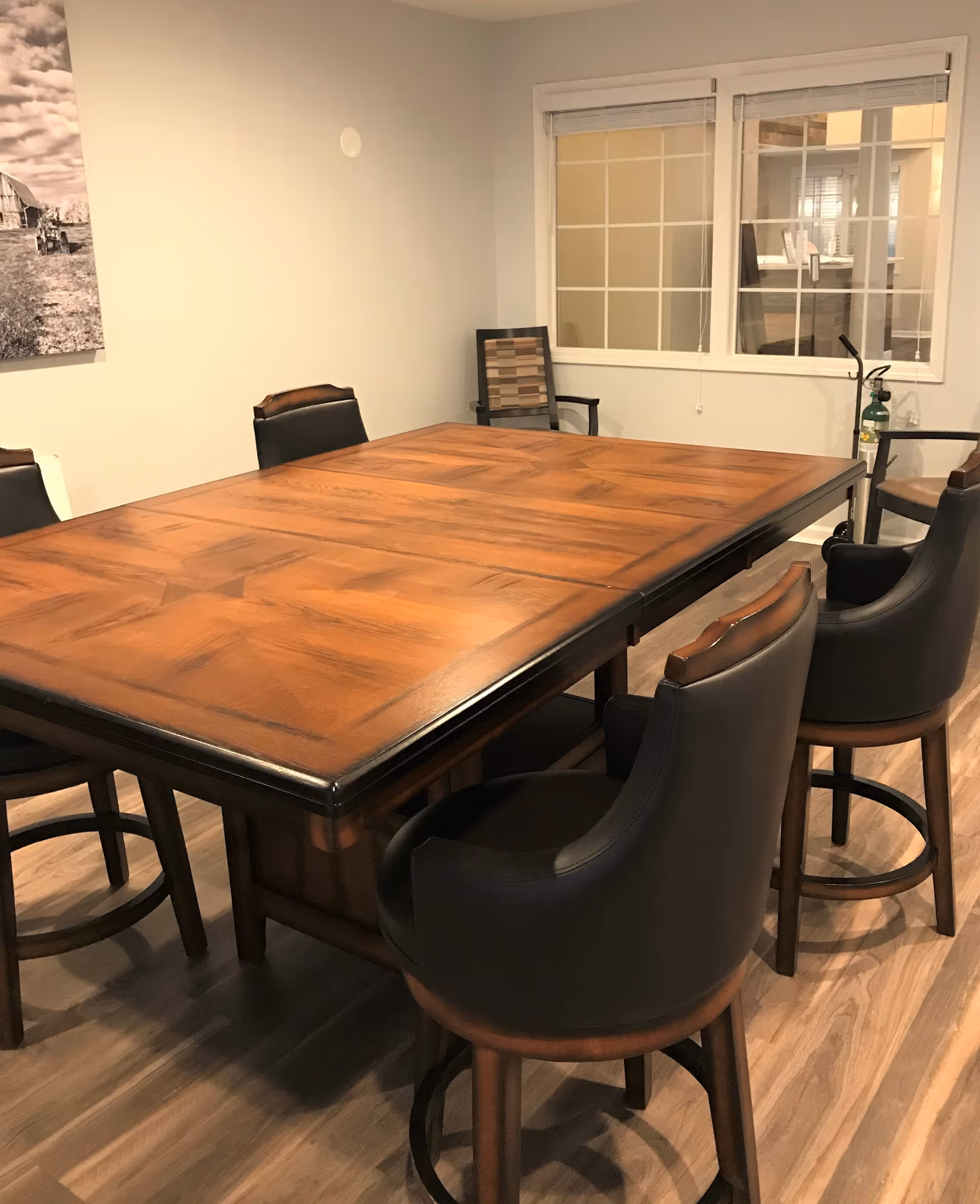 A wooden rectangular table surrounded by six cushioned chairs with dark leather upholstery in a room with light-colored walls and wood flooring. A window with white framing is visible on the wall behind the table, and a black and white photo hangs on the adjacent wall.