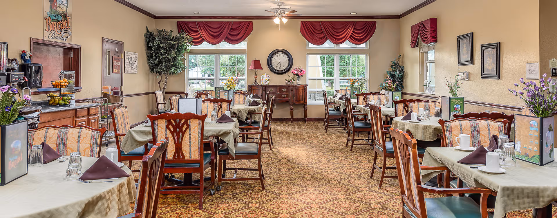 A well-lit dining room with multiple tables covered in beige tablecloths, each set with glasses, napkins, and cups. The room features wooden chairs with patterned upholstery, decorative plants, framed pictures on the walls, and large windows with red valances letting in natural light. A sideboard with a clock and flowers is visible at the far end of the room.