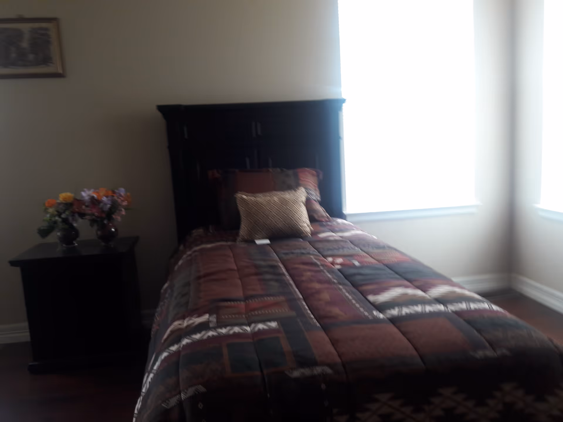 A bedroom with a single bed featuring a dark wooden headboard and a patterned bedspread in shades of brown, red, and green. There are two pillows on the bed, one larger and one smaller. To the left of the bed is a dark wooden nightstand with two vases of colorful flowers. The room has light-colored walls and two large windows letting in bright natural light.