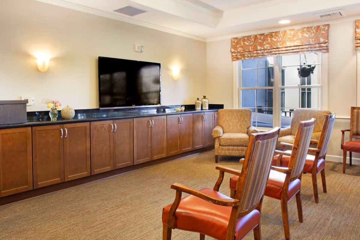 A cozy living room area with a large flat-screen TV mounted on the wall above a long wooden cabinet with multiple doors. The room has several chairs with wooden arms and red cushions arranged facing the TV, and two upholstered armchairs near a window with patterned valances. The walls are light-colored with two wall sconces providing soft lighting.